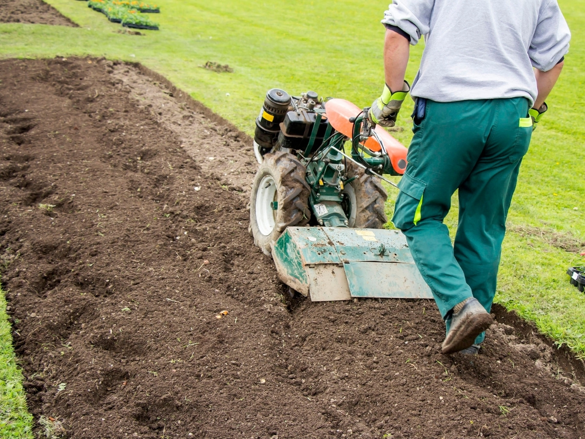 Person in Arbeitskleidung bedient motorisierten Bodenfräsen auf einem Feld mit frisch umgegrabener Erde