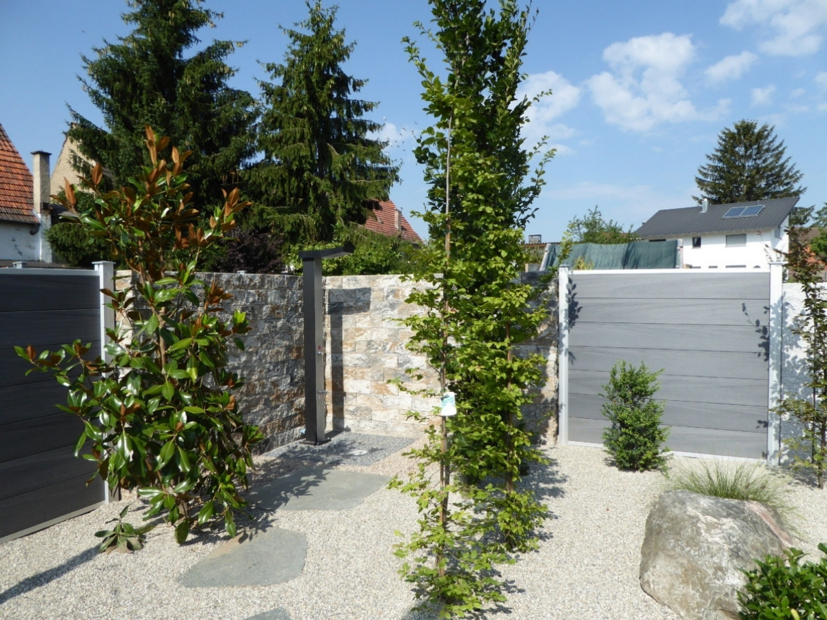 Garten mit Kiesboden, mehreren Bäumen, einer Steinmauer und grauen Holzzäunen unter blauem Himmel