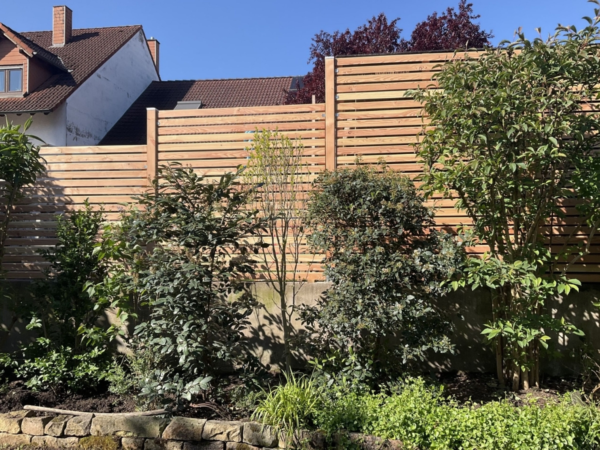 Holzzaun mit horizontalen Latten hinter einem Garten mit verschiedenen Sträuchern und Bäumen