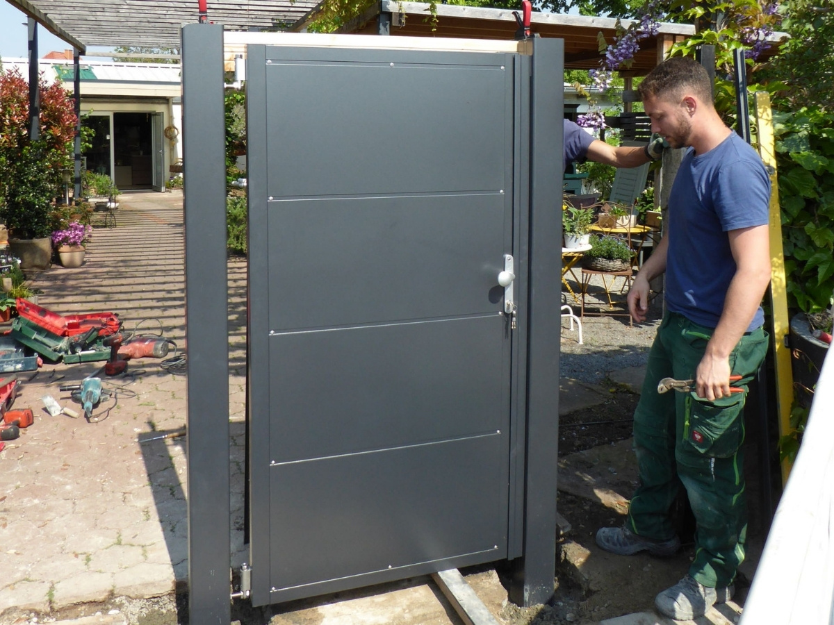 Mann in blauer T-Shirts und grüner Arbeitshose steht neben einem grauen Metalltor mit Griff und Schloss in einem Außenbereich mit Pflanzen und Werkzeugen