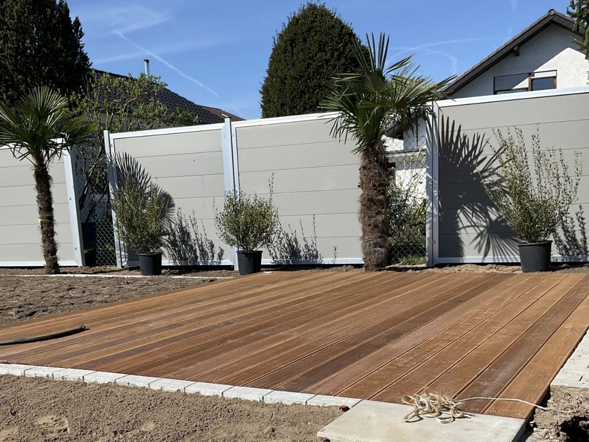 Holzterrasse vor einem weißen Sichtschutzzaun mit drei Palmen und zwei kleinen Büschen in Pflanzkübeln