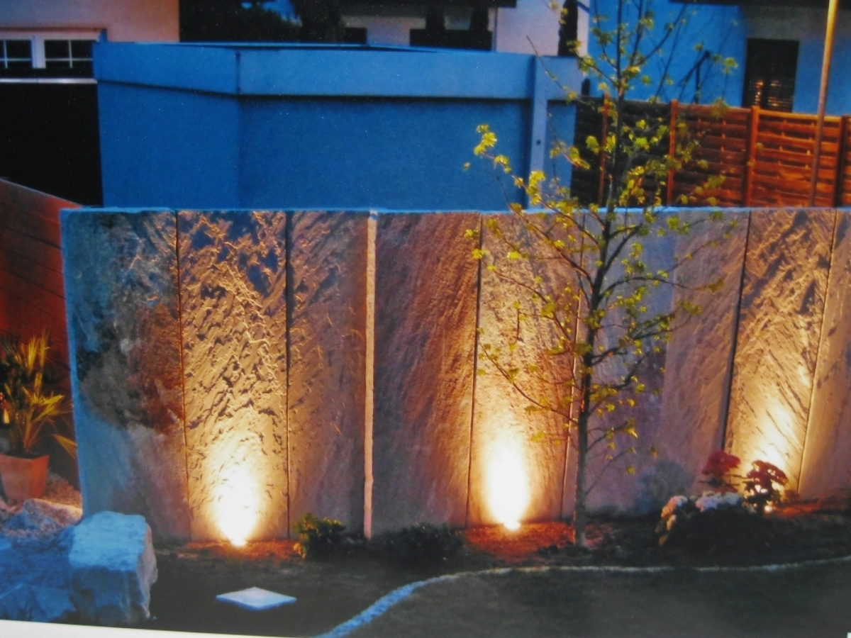 Beleuchtete Steinmauer mit vertikalen Platten und kleinen Pflanzen davor in einem Garten bei Dämmerung
